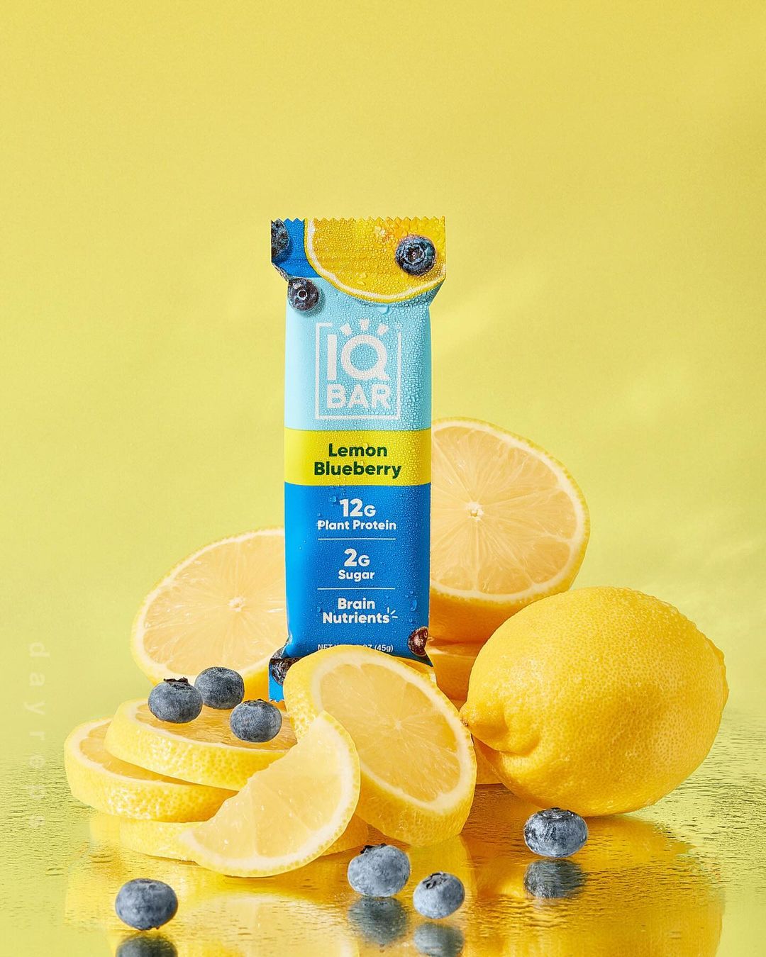 IQBAR Lemon Blueberry protein bar product shot with lemon slices and blueberries against a bright yellow background in studio.