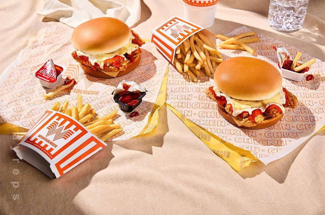 Two Whataburger chicken sandwiches and french fries arranged on logo-printed paper, with a cup of water in the background.