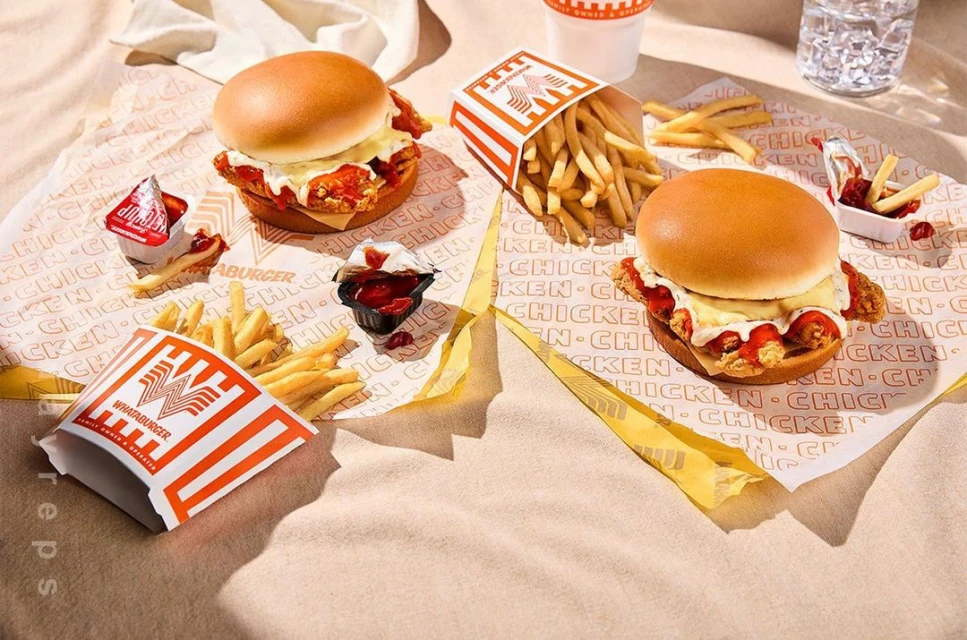 Two Whataburger chicken sandwiches and french fries arranged on logo-printed paper, with a cup of water in the background.