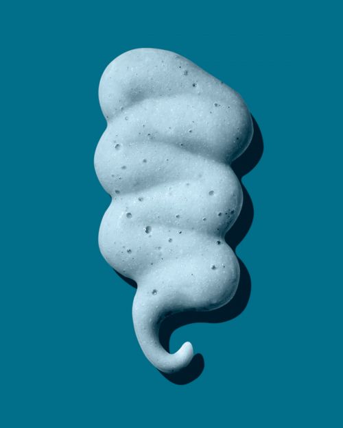 Close-up studio shot of a textured swirl of cleansing foam against a solid teal background, highlighting its smooth and airy consistency.