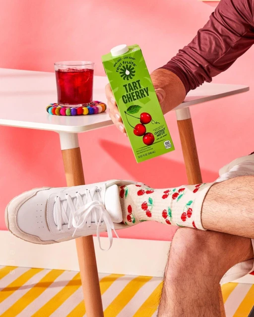 Hand holds tart cherry juice carton next to table with glass of juice, patterned coaster, and white sneakers on a striped surface.