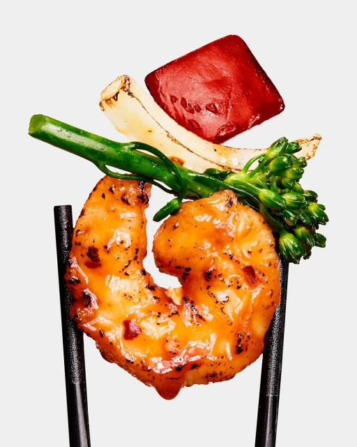 Close-up of grilled shrimp held by chopsticks, accompanied by grilled vegetables like red pepper, onion, and broccolini, against a plain background.