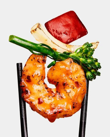 Close-up of grilled shrimp held by chopsticks, accompanied by grilled vegetables like red pepper, onion, and broccolini, against a plain background.
