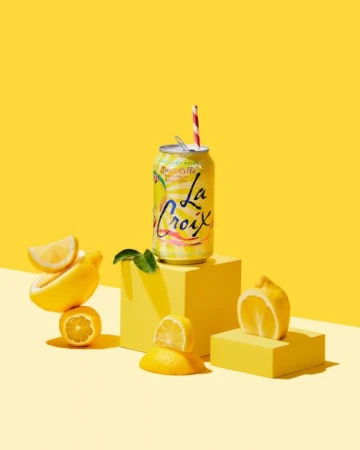La Croix sparkling water can with a striped straw, arranged with cut lemons on yellow geometric blocks against a yellow background.