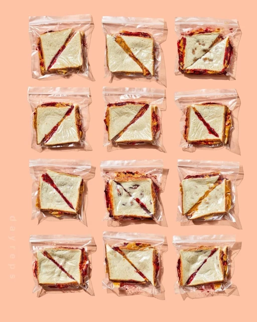 Eleven sealed plastic bags, each containing a diagonally-cut sandwich, arranged in rows against a light peach-colored background. Food photography.