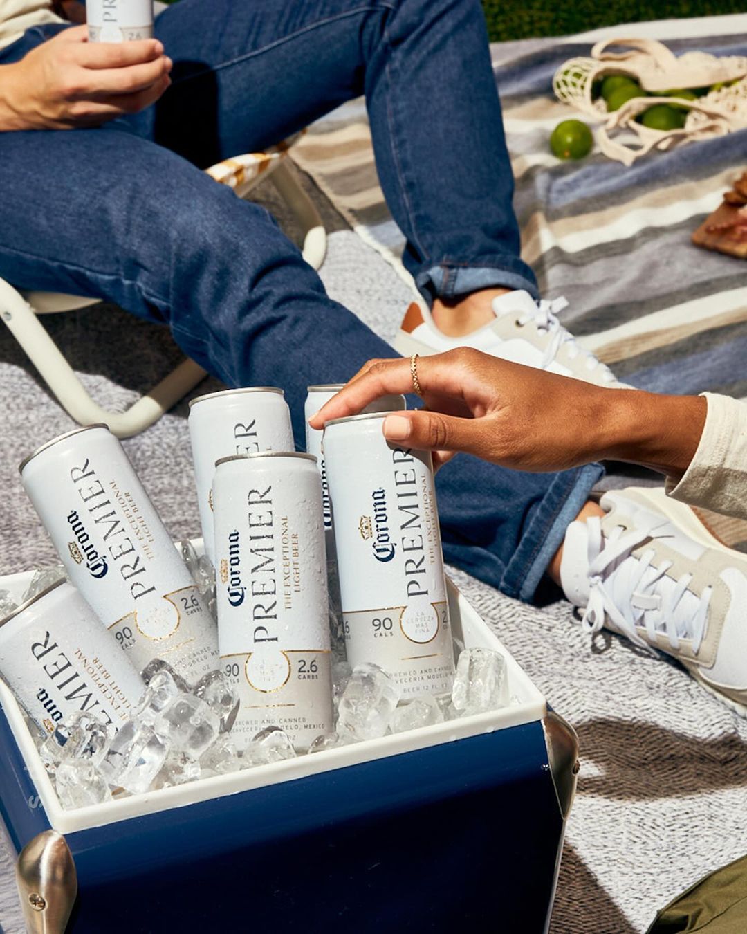 Two adults enjoy refreshing Corona Premier beer from an ice-filled cooler, sitting on a blanket outside. Limes and a tote bag are visible.