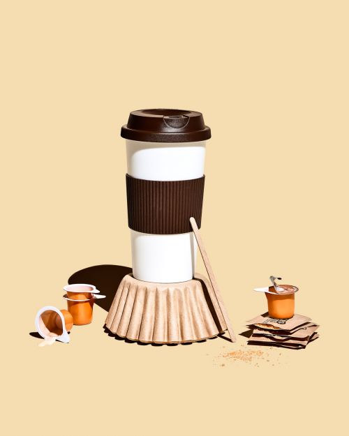 Still life shot of a white reusable coffee cup with brown lid and sleeve, with coffee pods and accessories against a light beige background.