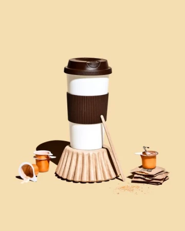 Still life shot of a white reusable coffee cup with brown lid and sleeve, with coffee pods and accessories against a light beige background.