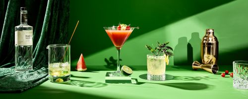 A vibrant still life showcases an assortment of colorful cocktails and ingredients against a bright green backdrop with dramatic shadows.