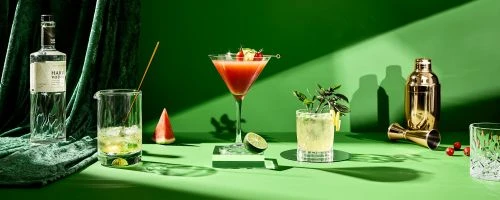 A vibrant still life showcases an assortment of colorful cocktails and ingredients against a bright green backdrop with dramatic shadows.