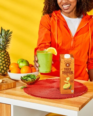 Cheerful woman in orange jacket holds green smoothie with fruit and juice box on a light wooden kitchen counter, bright yellow background.