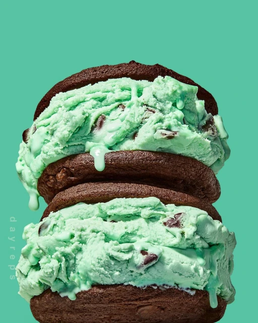 Close-up of a mint chocolate chip ice cream sandwich made with two chocolate cookies, set against a solid teal background.