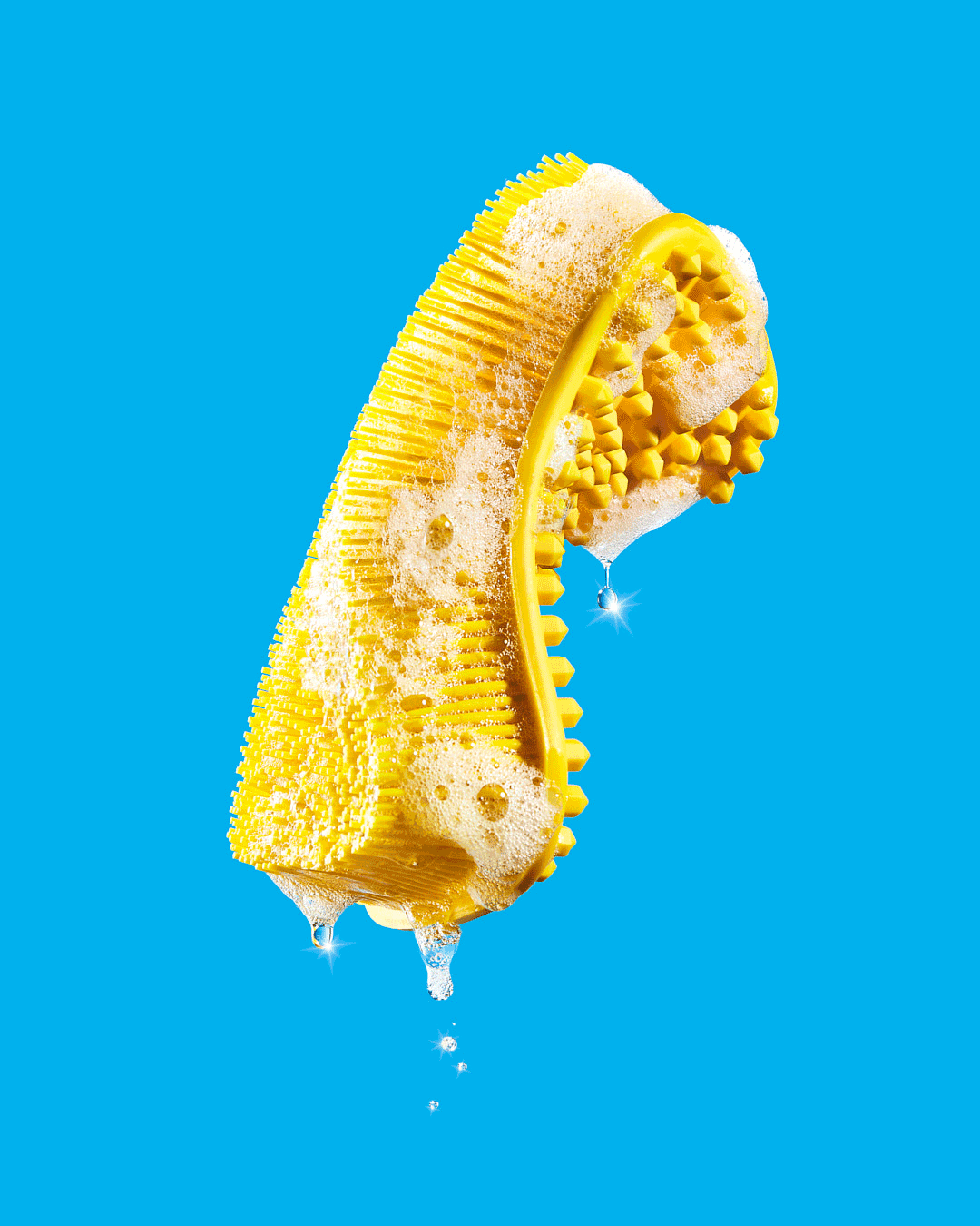 Yellow textured cleaning sponge covered in bubbles and dripping water on a bright blue background in a studio shot.