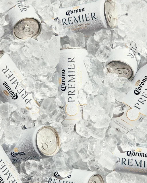 Overhead shot of white Corona Premier beer cans nestled in crushed ice, creating a refreshing, cool beverage scene.