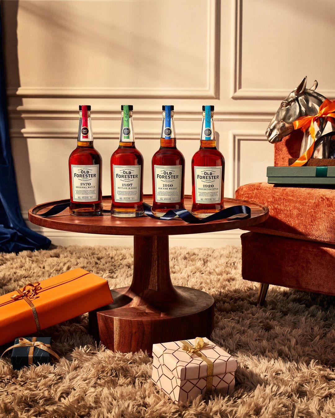 Four bottles of Old Forester bourbon whiskey are displayed on a wooden table, surrounded by gifts and a decorative horse head.