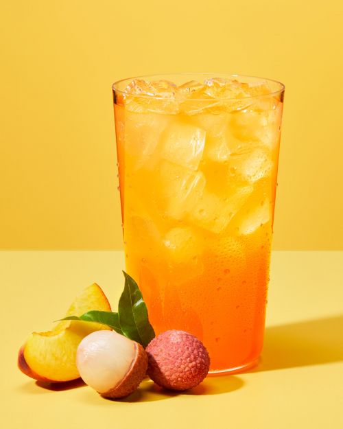 Tall glass filled with orange beverage, ice, peach slices, and lychee against a solid yellow backdrop in a brightly lit food still life.