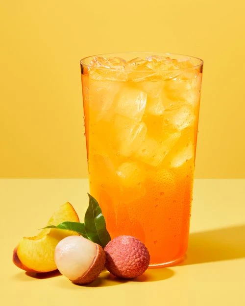 Tall glass filled with orange beverage, ice, peach slices, and lychee against a solid yellow backdrop in a brightly lit food still life.