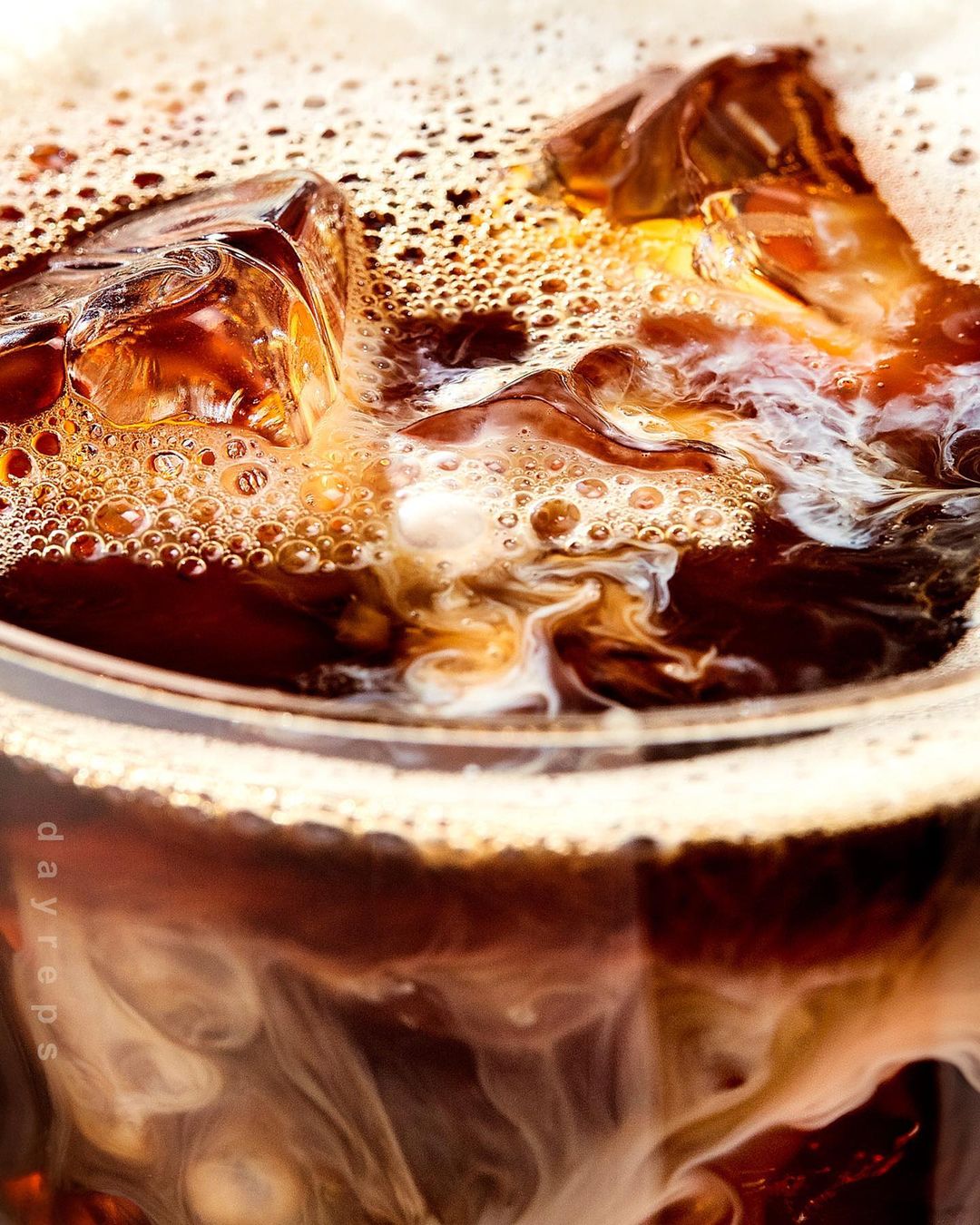 Close-up of iced coffee in a glass with ice cubes and cream swirling to create a textured, refreshing beverage drink.
