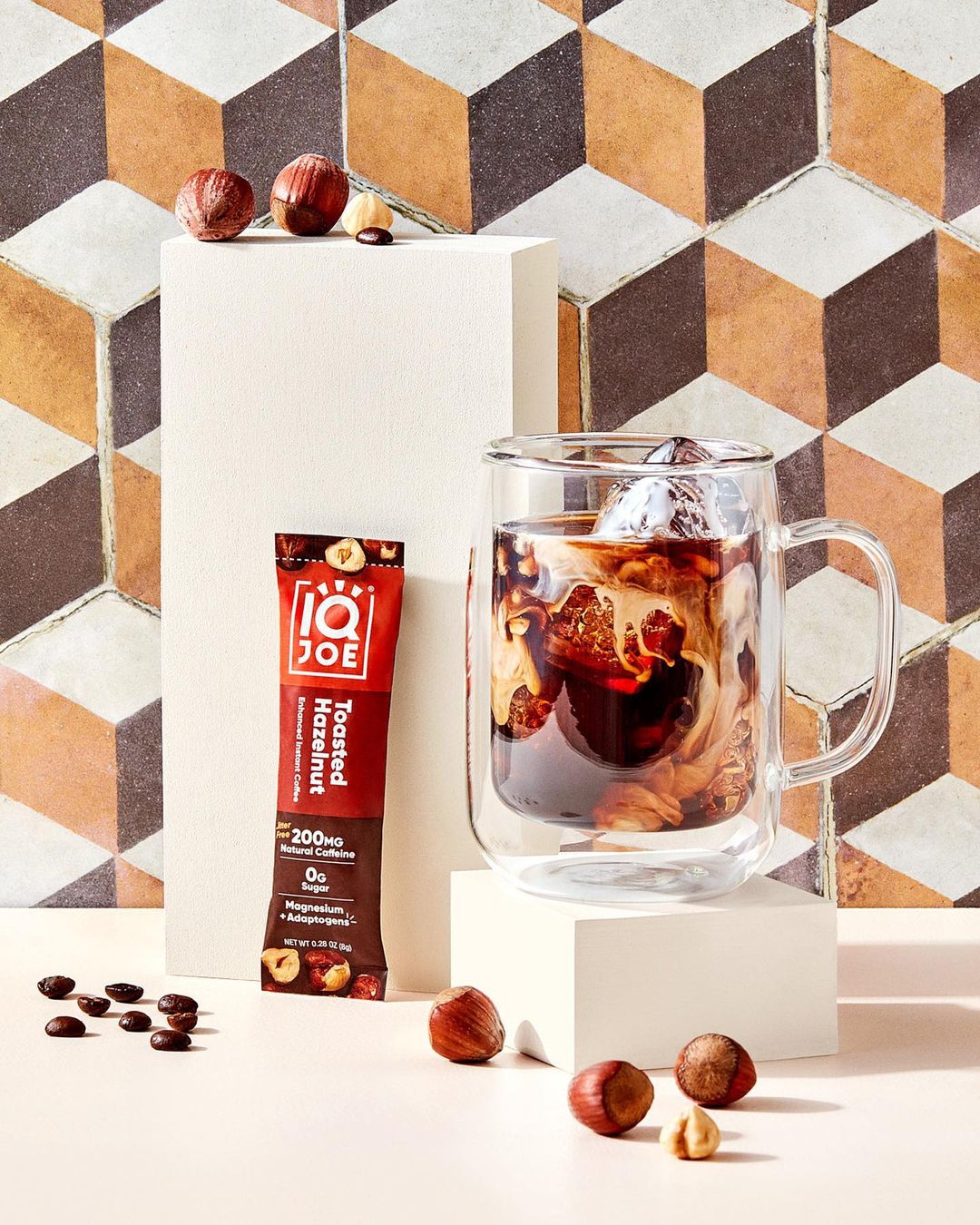 Coffee drink in clear glass mug with ice, hazelnuts, and IQJOE Toasted Hazelnut packet on cream-colored surfaces and a geometric tiled wall.