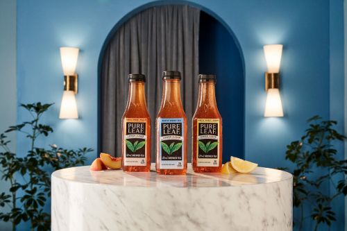 Three bottles of Pure Leaf iced tea are lined up on a marble table with peach and lemon slices, set against a blue wall with sconces.