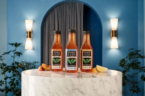 Three bottles of Pure Leaf iced tea are lined up on a marble table with peach and lemon slices, set against a blue wall with sconces.