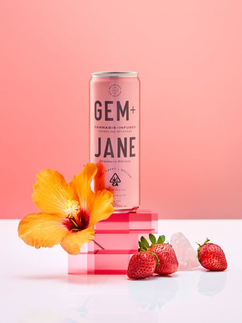 Pink cannabis-infused drink can with Gem+Jane branding, accented by a vibrant hibiscus, fresh strawberries, and clear pink block decor.