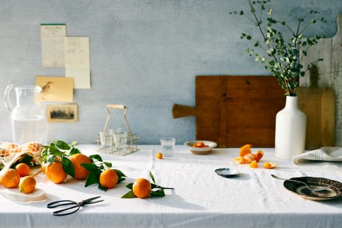 Still life on a white tablecloth featuring a glass pitcher, oranges with leaves, a white vase with greenery, and wooden cutting boards.