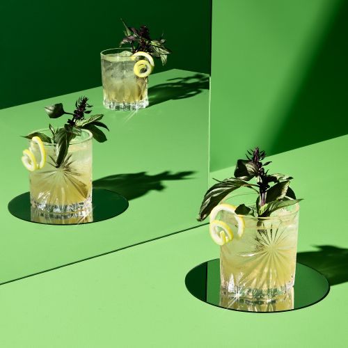 Three glasses of cocktails are garnished with lemon twists and basil, arranged on a green surface with matching backdrop, creating a vibrant, fresh scene.