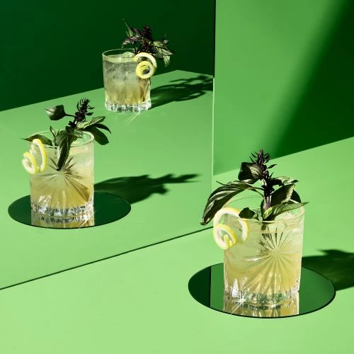 Three glasses of cocktails are garnished with lemon twists and basil, arranged on a green surface with matching backdrop, creating a vibrant, fresh scene.