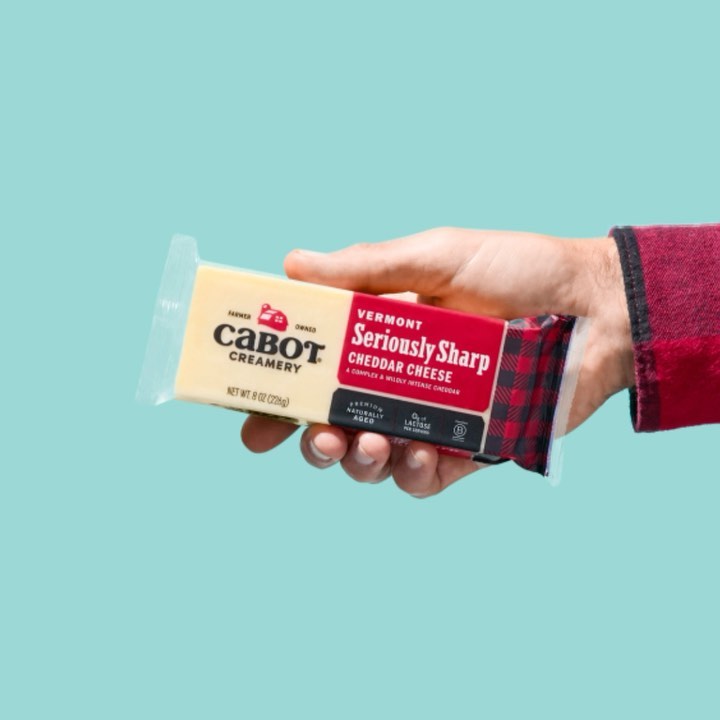 Person holds a package of Cabot Seriously Sharp Cheddar Cheese in front of a light blue background. Red plaid sleeve visible.