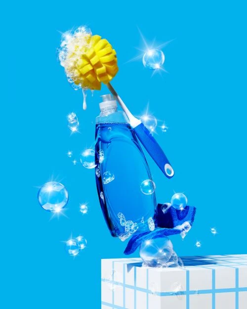 Still life composition of blue dish soap bottle, scrub brush, sponge, and floating bubbles against a bright turquoise background.