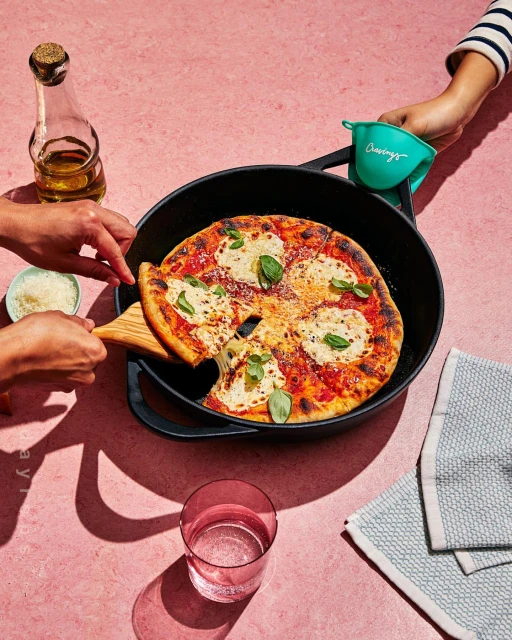 Overhead shot of a pizza in a cast-iron skillet with a person taking a slice, surrounded by oil, a water glass, and dishcloths.