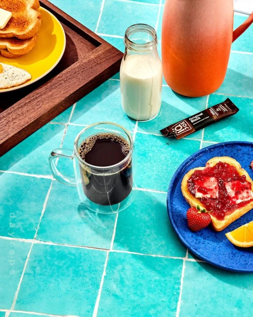 Overhead shot of breakfast scene: coffee, milk, toast with jam, egg, and orange slice on a turquoise tile surface.