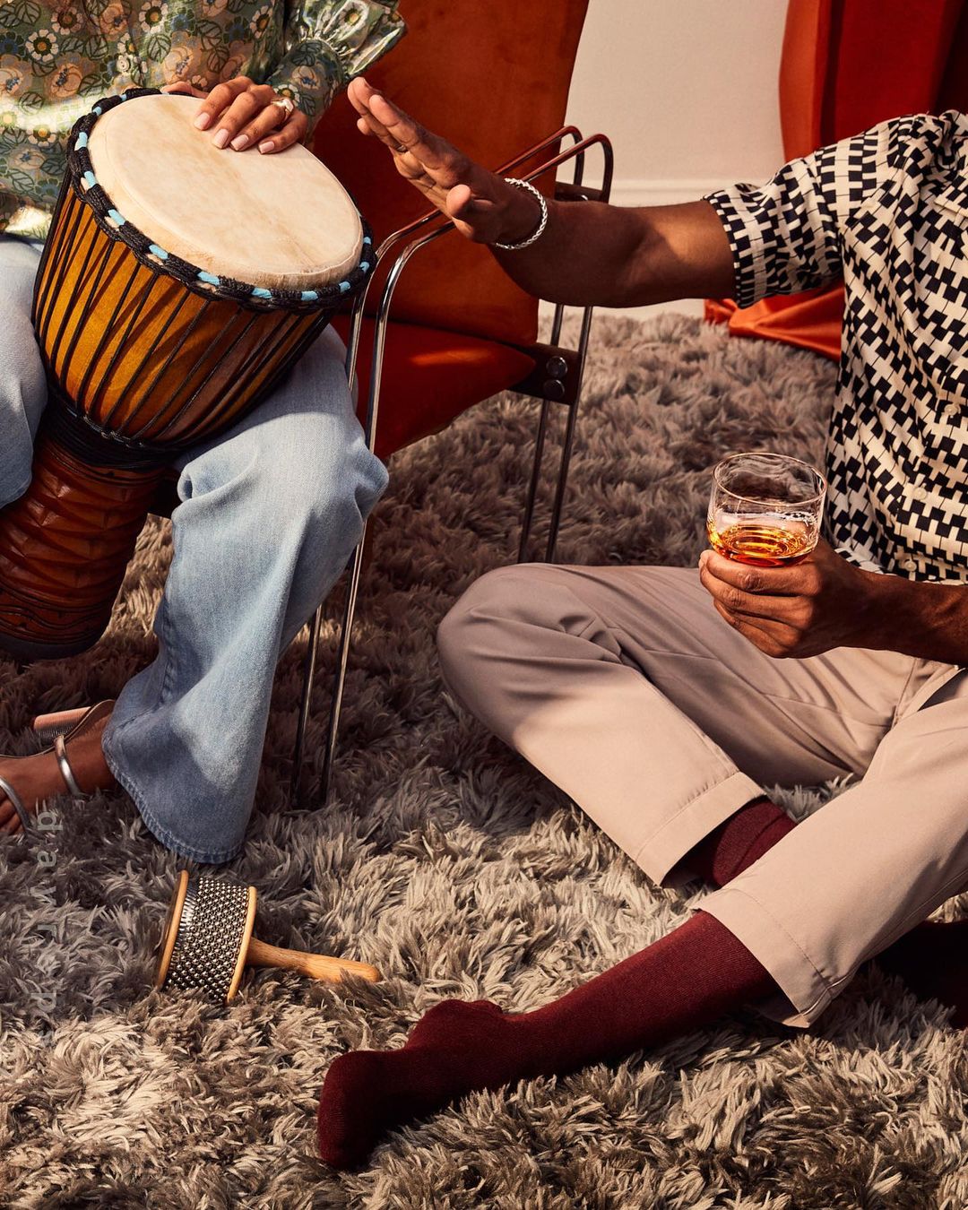 Two individuals are indoors, one playing a djembe drum, the other holding a glass, seated on a grey, furry rug next to an orange chair.
