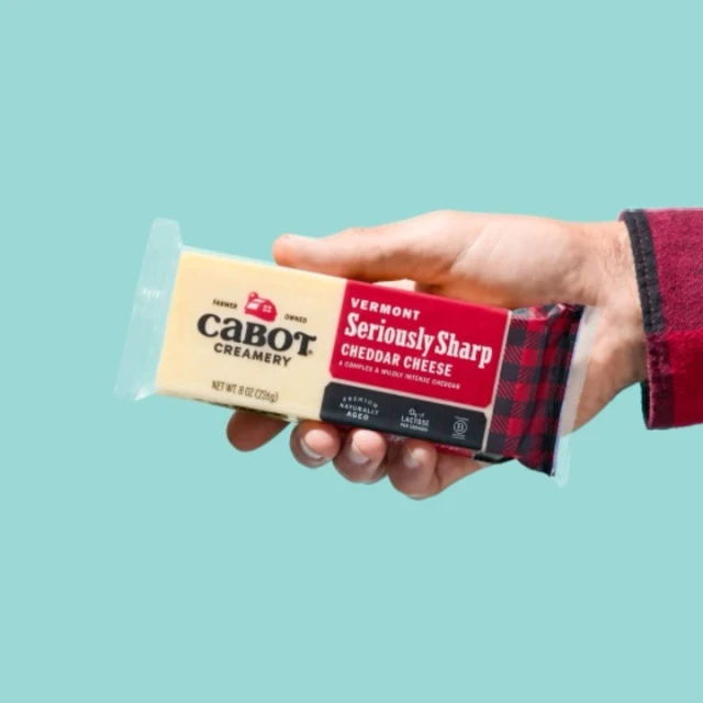 Hand holding a bar of Cabot Seriously Sharp Cheddar Cheese against a blue background, showcasing the product and its branding.
