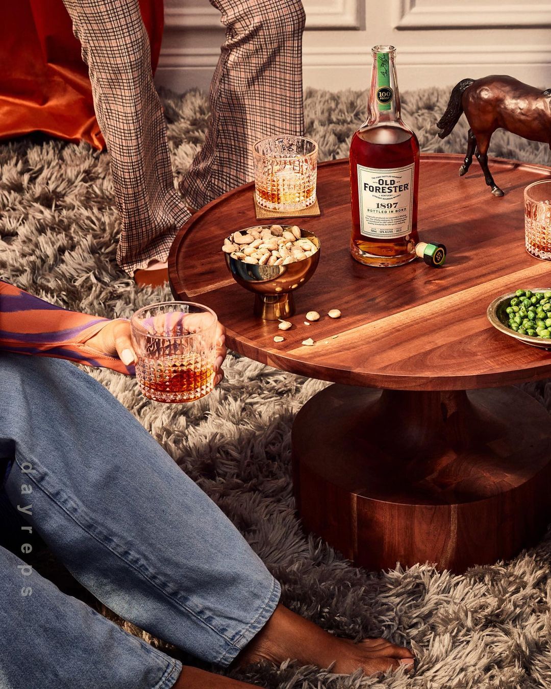 Indoor social gathering with people drinking whiskey, snack table with bottle of Old Forester, nuts, and peas, and a horse figure.