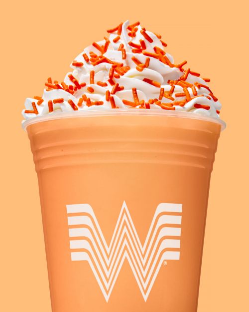 Close-up studio shot of a creamy orange milkshake topped with whipped cream and orange sprinkles against a matching orange background.