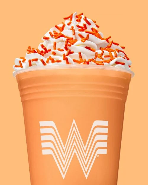 Close-up studio shot of a creamy orange milkshake topped with whipped cream and orange sprinkles against a matching orange background.