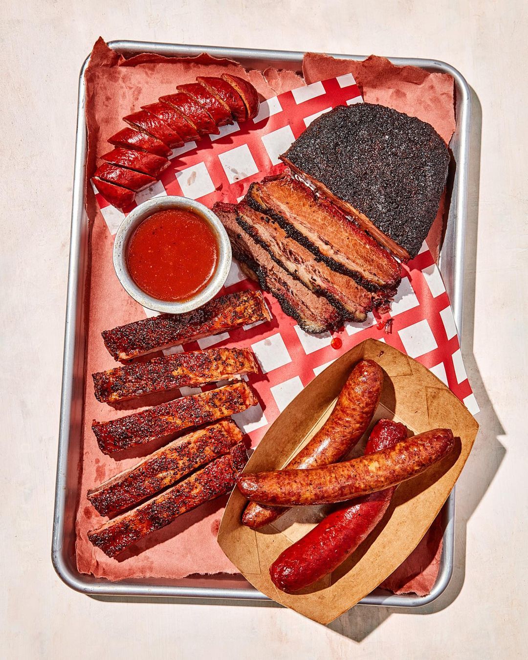 Overhead shot of a silver tray holding sliced sausage, brisket, ribs, a bowl of red sauce, and sausage links on a red and white checkered paper.