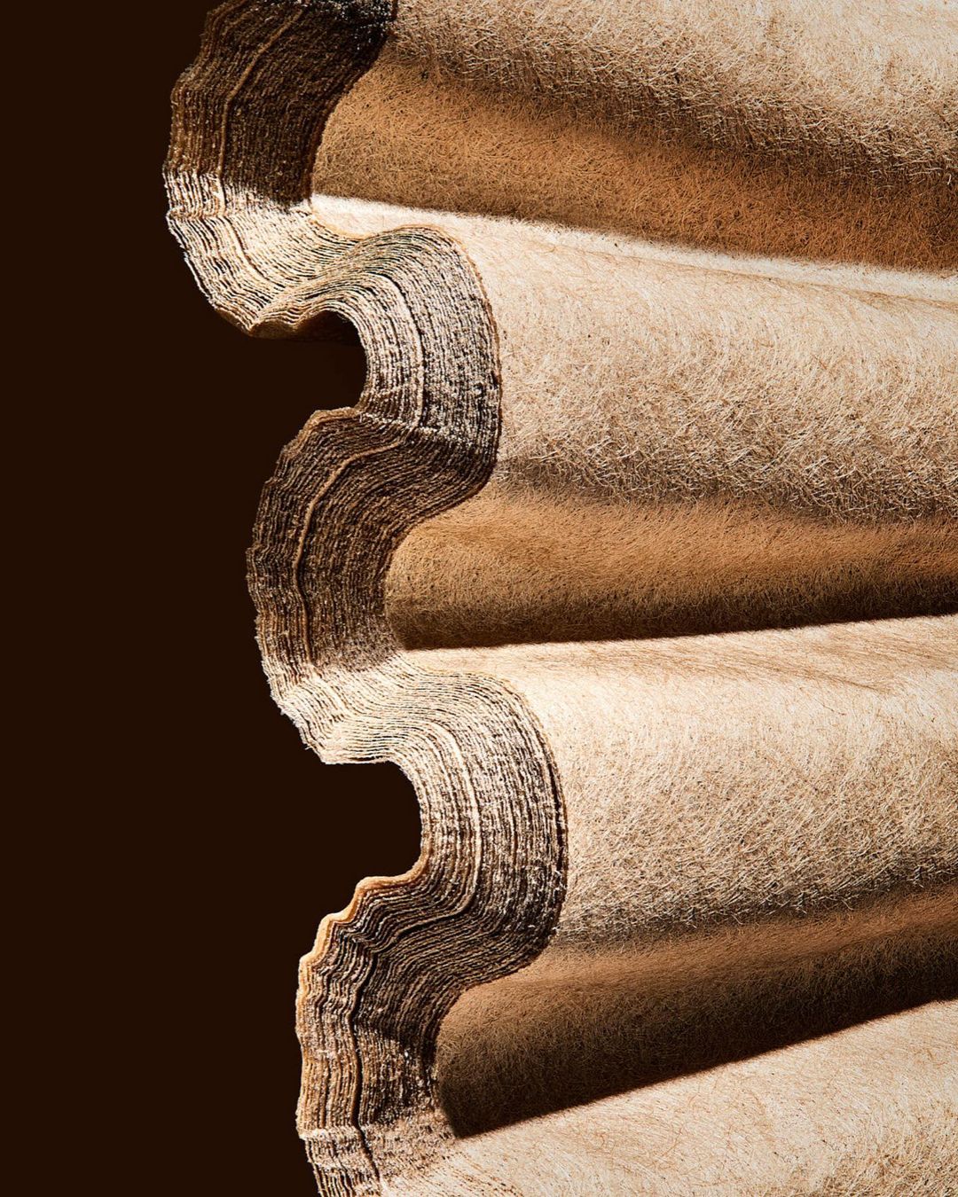 Close-up of stacked, corrugated beige material against a dark background, highlighting texture and repeating pattern.