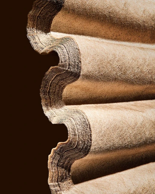 Close-up of stacked, corrugated beige material against a dark background, highlighting texture and repeating pattern.