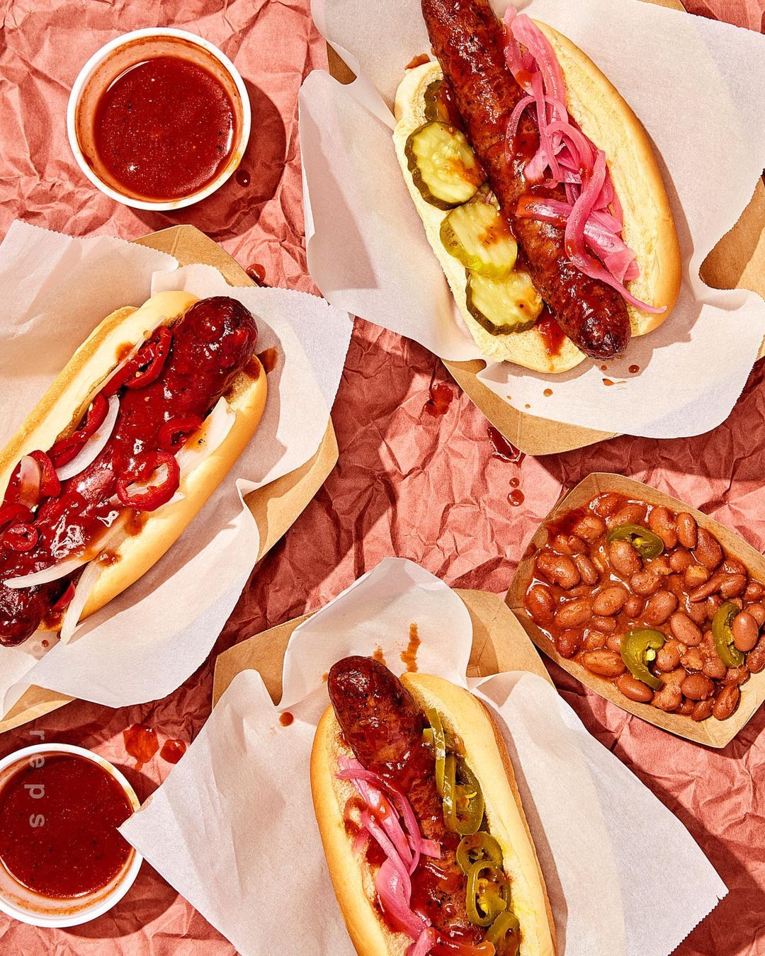 Overhead shot of three hot dogs with assorted toppings on white paper with a side of baked beans and red sauce on maroon paper.