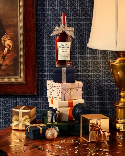 Festive still life with an Old Forester bourbon bottle atop gift boxes, complemented by glittery ornaments and ribbons on a wood surface.