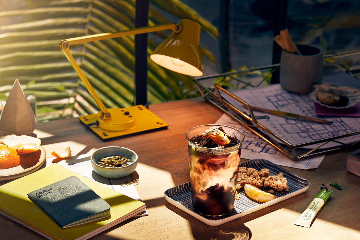 A glass of iced coffee with cream sits on a wooden desk beside a yellow lamp, notebooks, and snacks in an inviting, sunlit setting.