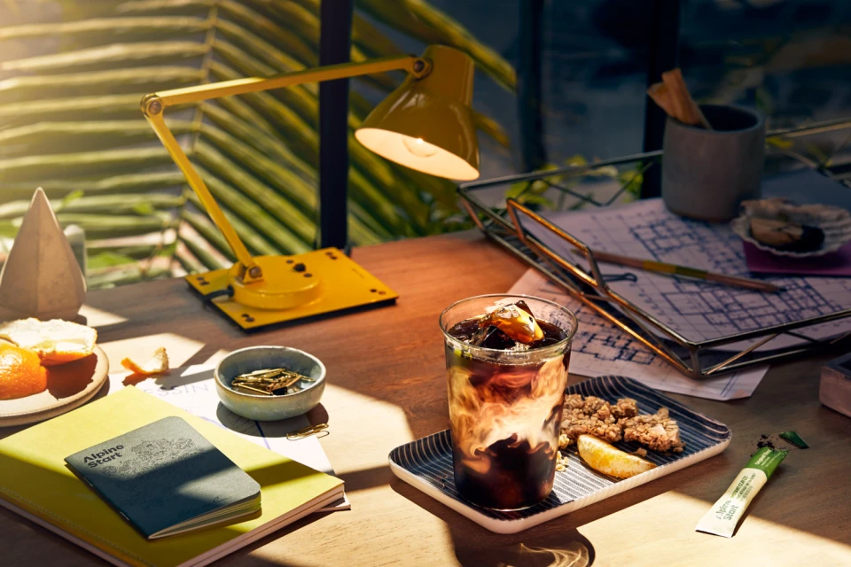A glass of iced coffee with cream sits on a wooden desk beside a yellow lamp, notebooks, and snacks in an inviting, sunlit setting.