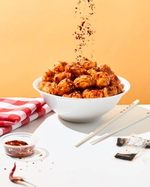 Bowl of glazed chicken pieces, chopsticks, and a red-and-white checkered cloth on a white surface against an orange background.