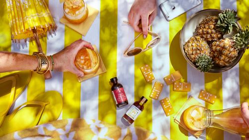 Overhead shot of pineapple, glasses, cocktails, yellow dominoes, umbrella and hands over a yellow and white striped surface.