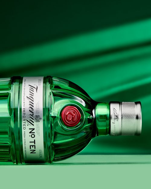 Tanqueray No. Ten gin bottle lying horizontally on a green surface, label prominently displayed, against a gradient green backdrop.