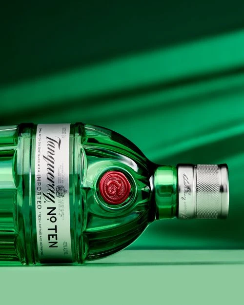 Tanqueray No. Ten gin bottle lying horizontally on a green surface, label prominently displayed, against a gradient green backdrop.
