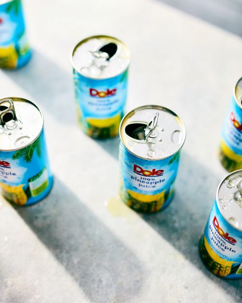 Group of opened Dole pineapple juice cans arranged on a textured surface, captured with bright lighting and a close-up perspective.